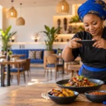 Restaurant owner in a blue headwrap photographing a plate of African dishes with her phone